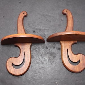 May include: A pair of matching wall-mounted wooden shelves, each featuring a curved, scroll-like support structure with a cutout design and a small hole at the top for hanging. The wood has a medium-brown finish with visible grain patterns.