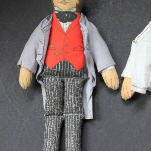 May include: A vintage fabric doll dressed in a gray jacket, red vest, and black and white striped pants. The doll has a painted face with brown hair and a black bow tie.