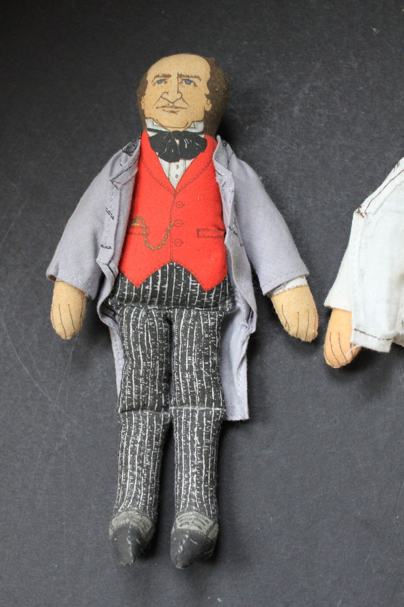 May include: A vintage fabric doll dressed in a gray jacket, red vest, and black and white striped pants. The doll has a painted face with brown hair and a black bow tie.
