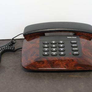 May include: A RadioShack landline telephone featuring a faux wood grain finish on the base and a black handset. The device includes a standard numeric keypad with black buttons, a coiled black cord, and function keys labeled Redial, Mute, Flash, and Hold.