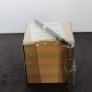 May include: A square memo cube with alternating orange and yellow paper sheets, wrapped in clear plastic. A black pen rests diagonally across the top surface, which features a white label with the text MEMO CUBE and OLYMPIA SALES ENFIELD.