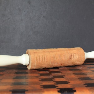 May include: A wooden rolling pin with white handles. The rolling pin has a carved design on the surface. It is lying on a wooden cutting board.
