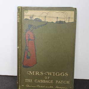 May include: A hardcover book titled Mrs. Wiggs of the Cabbage Patch by Alice Caldwell Hegan. The olive green cover features a graphic illustration of a person in a red dress standing near a fence, with a landscape scene above the title text.