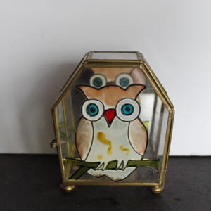 May include: A small, hexagonal glass and brass trinket box featuring a hand-painted owl design. The owl has large blue eyes, a red beak, and is perched on a green branch. The box has a hinged lid and sits on four small brass feet.