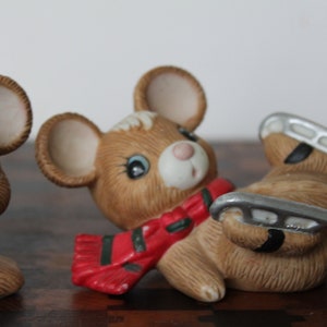 May include: Three ceramic mouse figurines with painted details. One mouse is wearing a red and green scarf, another is wearing a red and green scarf and ice skates, and the third mouse is wearing a red and green scarf.