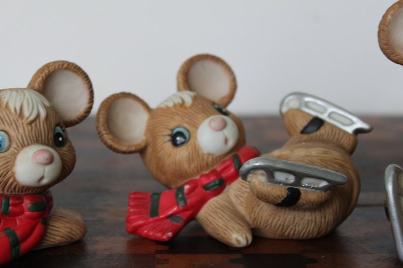May include: Three ceramic mouse figurines with painted details. One mouse is wearing a red and green scarf, another is wearing a red and green scarf and ice skates, and the third mouse is wearing a red and green scarf.