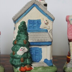 May include: A white ceramic house with a blue roof and blue windows. There is a green tree in front of the house and a ghost on the roof. The house has a blue door with the number 1212 on it.