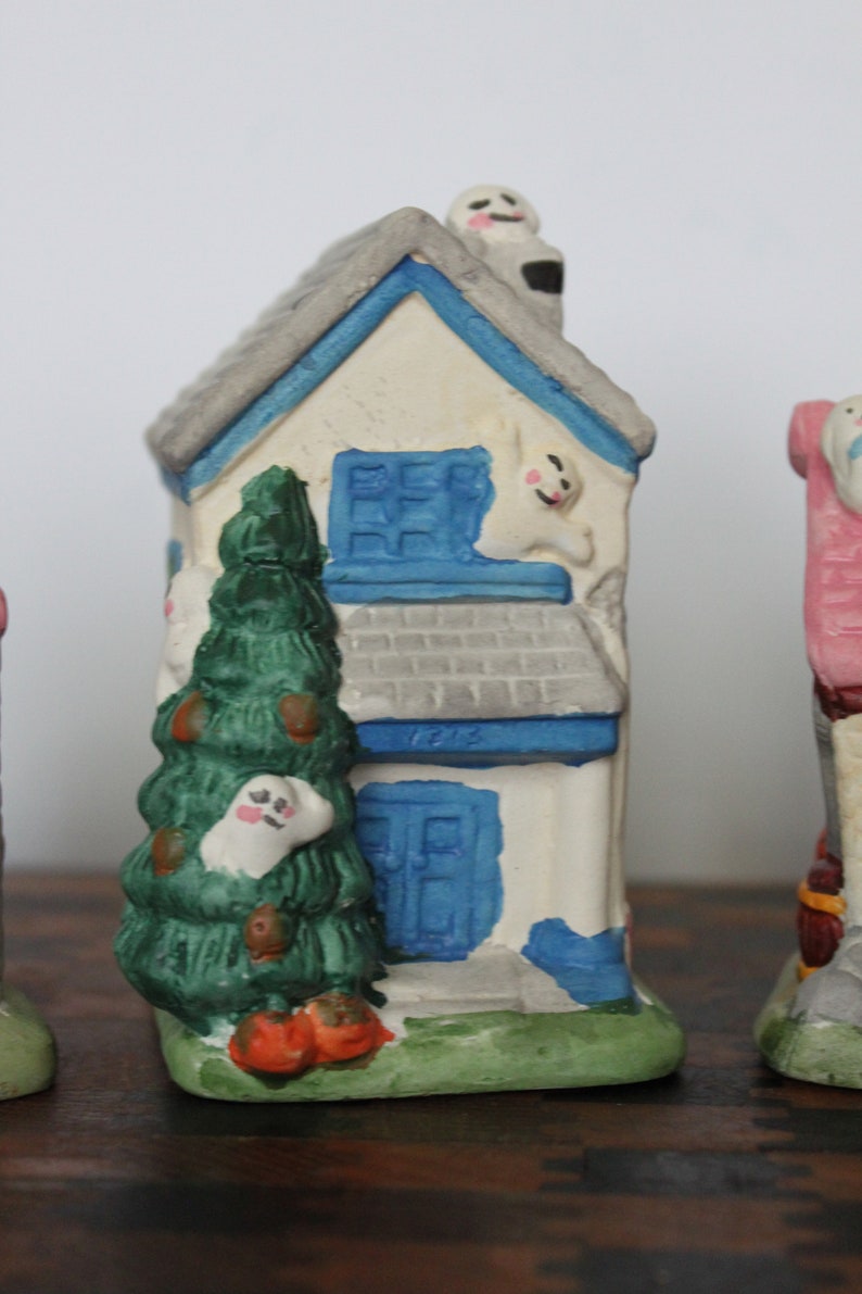 May include: A white ceramic house with a blue roof and blue windows. There is a green tree in front of the house and a ghost on the roof. The house has a blue door with the number 1212 on it.