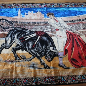 May include: A decorative tapestry depicting a bullfight scene. A black bull charges towards a matador in a white suit and red cape. The background shows a stadium and blue sky, with a decorative border.