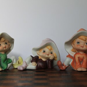 May include: Three ceramic figurines of whimsical elves. Each elf wears a large hat and colorful clothing in shades of green, pink, and orange. The elves have rosy cheeks and are posed in relaxed positions.