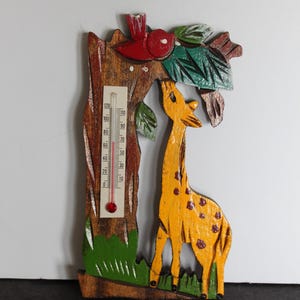 May include: Wooden thermometer shaped like a tree with a giraffe. The tree is brown with green leaves and grass. A red bird sits on top. The giraffe is yellow with brown spots. The thermometer reads in Fahrenheit.