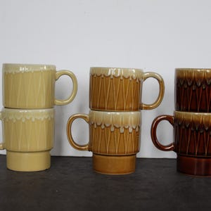 May include: Stacked ceramic mugs in shades of yellow, brown, and dark brown. The mugs have a textured design and a handle. The mugs are stacked on top of each other.