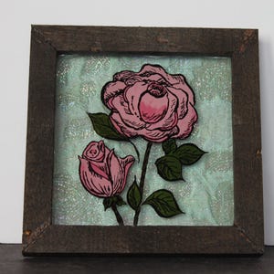 May include: Square framed artwork featuring a pink rose and bud with green leaves against a textured, light blue background. The artwork is enclosed in a dark brown wooden frame, creating a rustic aesthetic. The rose is hand-drawn with black outlines.