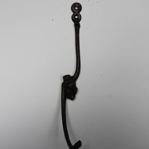 May include: A dark brown, cast iron wall hook. The hook has a decorative design at the base and two mounting holes at the top. The hook curves downward, perfect for hanging coats or other items.