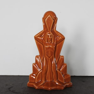 May include: A brown ceramic art deco figurine. The figurine depicts a stylized human figure with arms raised, set against a geometric backdrop. The glossy finish and warm color give it a vintage feel.