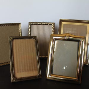 May include: Five vintage picture frames in various gold tones. The frames have different shapes and decorative details, including beaded edges and ornate curves. The frames are empty, ready to display photos or artwork.