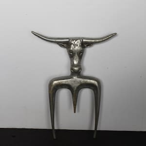 May include: A silver-colored metal serving fork with a bull head design. The fork has three tines and a long handle. The bull's head is detailed with horns and facial features. The object is likely used for serving food.