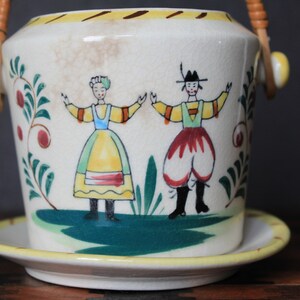May include: A white ceramic planter with a yellow rim and a brown woven handle. The planter features a painted scene of two figures in traditional clothing dancing in a green field with red berries and green leaves.