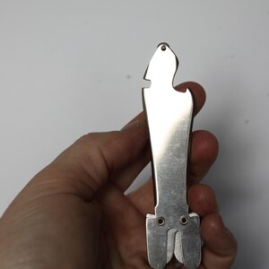 K&ouml;nnte beinhalten: Ein silberfarbener Flaschen&ouml;ffner mit einem einzigartigen, l&auml;nglichen Design. Das Werkzeug verf&uuml;gt &uuml;ber einen Flaschen&ouml;ffner, einen Kapselheber und ein kleines Loch oben. Das Werkzeug wird in einer Hand vor wei&szlig;em Hintergrund gehalten.