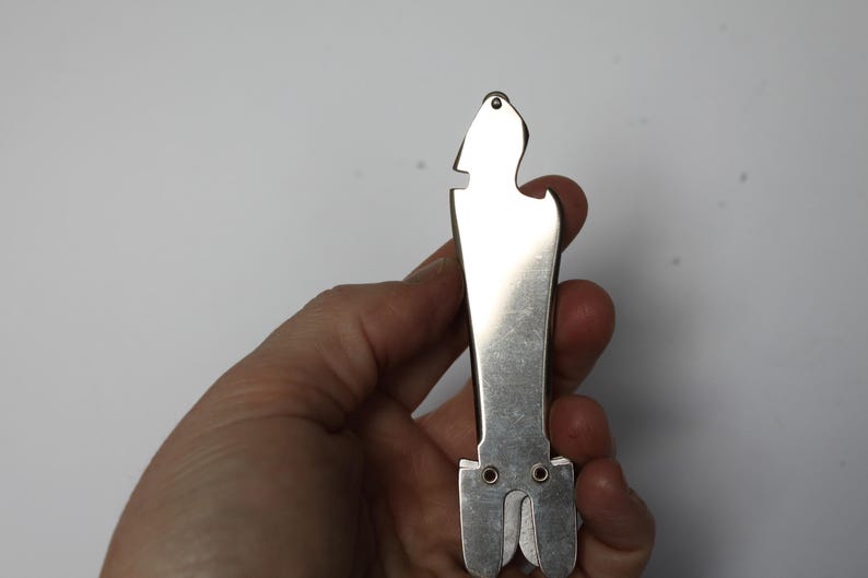 K&ouml;nnte beinhalten: Ein silberfarbener Flaschen&ouml;ffner mit einem einzigartigen, l&auml;nglichen Design. Das Werkzeug verf&uuml;gt &uuml;ber einen Flaschen&ouml;ffner, einen Kapselheber und ein kleines Loch oben. Das Werkzeug wird in einer Hand vor wei&szlig;em Hintergrund gehalten.