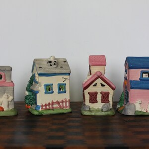 May include: Set of five miniature ceramic houses with Halloween themes. The houses are painted in various colors, including pink, blue, white, and gray. Each house has a unique design, including a church, a schoolhouse, and a house with a fence. The houses are decorated with ghosts and other Halloween-themed details.