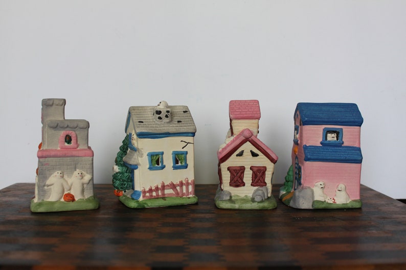 May include: Set of five miniature ceramic houses with Halloween themes. The houses are painted in various colors, including pink, blue, white, and gray. Each house has a unique design, including a church, a schoolhouse, and a house with a fence. The houses are decorated with ghosts and other Halloween-themed details.