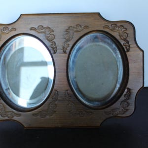 May include: A wooden double photo frame with two oval openings. The frame has a light brown finish and decorative scrollwork at the corners and between the openings. The glass in the openings reflects the light.
