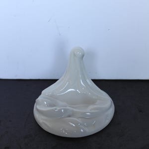 May include: A white ceramic decorative piece with a unique, abstract design. The object has a rounded base with a textured, flowing shape that rises to a central point. The piece is smooth and glossy.