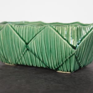 May include: A rectangular, green ceramic planter with a woven basket design. The planter has a textured surface with vertical lines and a scalloped top edge. The planter is approximately 10 inches long, 5 inches wide, and 4 inches tall.