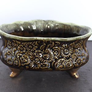 May include: An oval ceramic planter with three small feet, featuring a dark brown glossy glaze and a raised relief pattern of flowers and swirling foliage. The scalloped rim is finished with a lighter, muted green drip glaze effect.