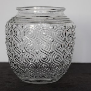 May include: Clear glass jar with a wide opening and a decorative pattern of swirling lines and dots. The jar has a rounded body and a slightly flared rim. The glass is transparent, allowing light to pass through.