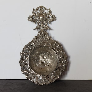 May include: Ornate silver tea strainer with a decorative handle and a detailed, pierced bowl. The bowl features a small, engraved scene. The strainer has an intricate, floral design around the edges and handle, adding to its antique aesthetic.