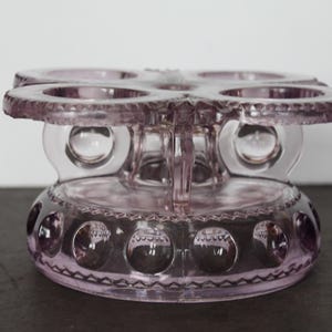 May include: A vintage, lavender-colored glass serving piece. It features a circular base with decorative circles, a central stem, and a top with four circular openings. The piece has a delicate, ornate design.