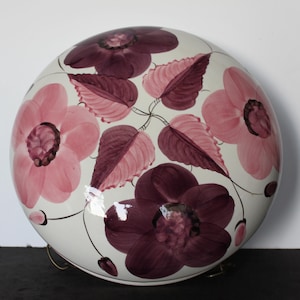 May include: A round, white ceramic decorative piece with a floral design. The flowers are in shades of pink and burgundy, with matching leaves and stems. The piece has a glossy finish and is likely a decorative bowl or lid.