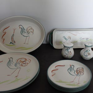 May include: A set of white ceramic dishes with a green rim and a rooster design. The set includes a large oval serving dish, two smaller round plates, a rectangular serving tray, and two salt and pepper shakers.