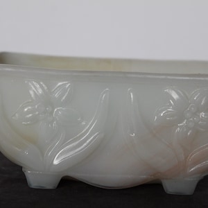 May include: A rectangular, off-white planter with a floral design. The planter has a raised floral pattern on the sides and sits on three small feet. The overall shape is a rectangle with rounded corners.