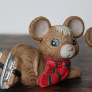 May include: Two brown ceramic mice figurines wearing red and green scarves. The mice are wearing silver ice skates and are sitting on a wooden surface.