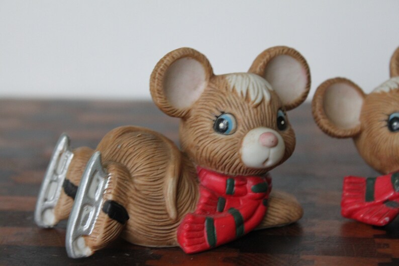 May include: Two brown ceramic mice figurines wearing red and green scarves. The mice are wearing silver ice skates and are sitting on a wooden surface.