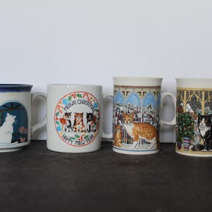 May include: Four ceramic mugs with cat-themed designs. One mug features a white cat in a window, another has a "Meowy Christmas" design with cats, and the others depict cats in decorative settings. Each mug has a white handle.