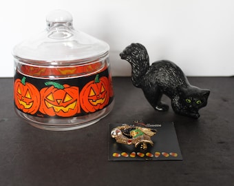 May include: A clear glass jar with a lid, decorated with orange jack-o'-lanterns. A black cat figurine and a Halloween-themed pin are also present. The pin features a bell and candy corn. Halloween theme.
