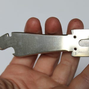 K&ouml;nnte beinhalten: Ein silbernes Metall-Multitool, das in einer Hand gehalten wird. Das Werkzeug hat einen Flaschen&ouml;ffner, einen Korkenzieher und eine kleine Klinge. Der Text "MADE IN GERMANY" ist auf dem Werkzeug eingestanzt. Das Werkzeug dient zum &Ouml;ffnen von Flaschen und zum Entfernen von Korken.