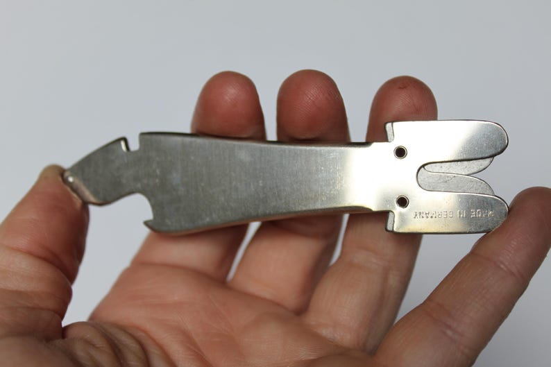 K&ouml;nnte beinhalten: Ein silbernes Metall-Multitool, das in einer Hand gehalten wird. Das Werkzeug hat einen Flaschen&ouml;ffner, einen Korkenzieher und eine kleine Klinge. Der Text "MADE IN GERMANY" ist auf dem Werkzeug eingestanzt. Das Werkzeug dient zum &Ouml;ffnen von Flaschen und zum Entfernen von Korken.