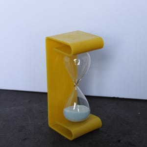 May include: A yellow hourglass with a C-shaped frame made of a smooth, solid material. The hourglass contains clear glass bulbs filled with fine, light blue sand, with the majority of the sand currently settled in the bottom bulb of the device.