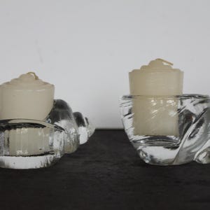 May include: Two clear glass seashell-shaped candle holders, each holding a white candle. The candle holders have a textured, swirling design. The candles are cylindrical with a small wick.