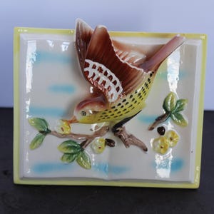 May include: A square ceramic wall plaque featuring a three-dimensional bird with brown wings and a yellow speckled body perched on a branch. The background is white with light blue horizontal accents, framed by a yellow border with small flowers.