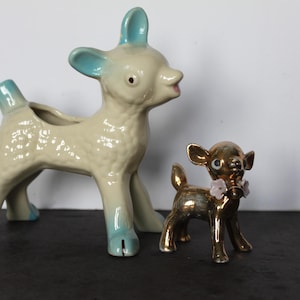 May include: Two ceramic deer figurines. One is a large, off-white planter with blue accents, and the other is a small, gold-colored deer with a floral collar. The deer are standing on a dark surface.