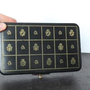 May include: A black rectangular box with a gold grid pattern and gold crest and knight designs. The box is held by a hand and has a gold clasp.