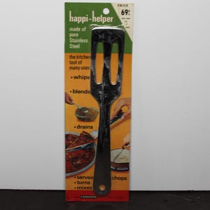 May include: A black stainless steel slotted kitchen spatula labeled happi-helper on its original cardboard packaging. The packaging features orange and green sections with text listing uses like whips, blends, drains, serves, turns, and mixes.