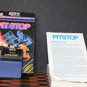 May include: A blue game cartridge for the Commodore 64 computer, labeled "PITSTOP™" and "Cartridge for COMMODORE 64". The cartridge is next to a box with a black and red illustration of a race car and the text "PIT STOP" in large white letters. The box also has the text "EPYX presents PITSTOP™" in white letters.