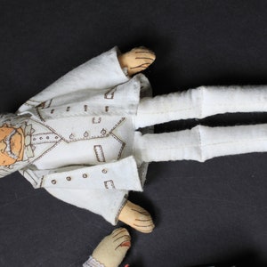 May include: A white fabric doll with a detailed face and a white suit. The doll is lying on a dark surface.